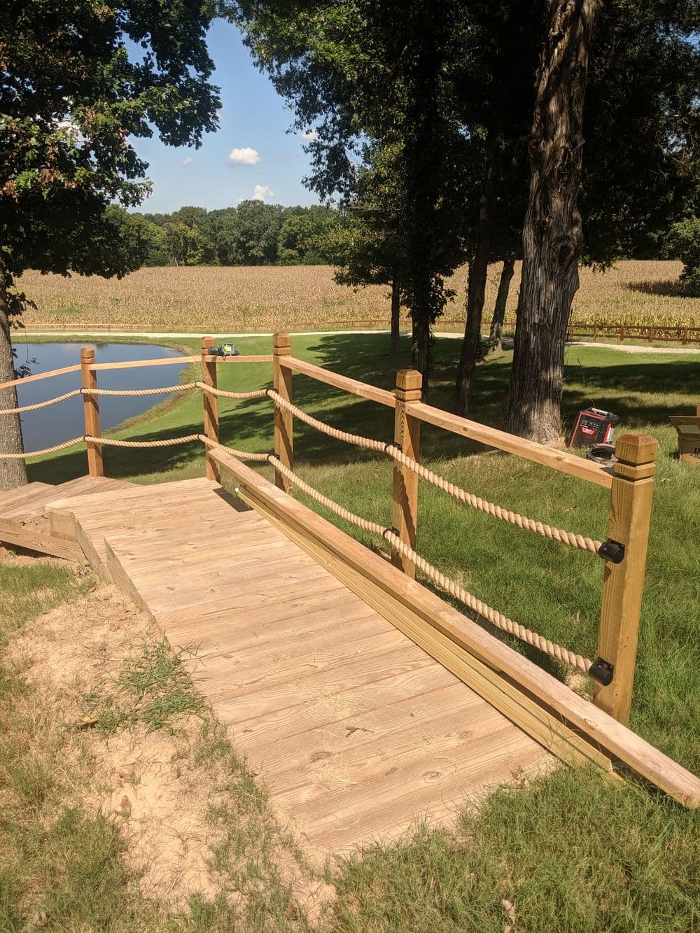 Wooden bridge with rope railing over a grassy area near a pond and cornfield in the background.
