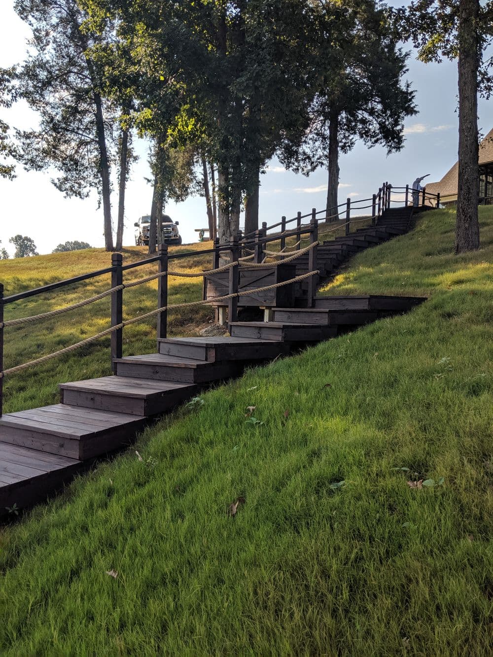 Wooden steps lead up a grassy hill; trees and a car are visible in the background.