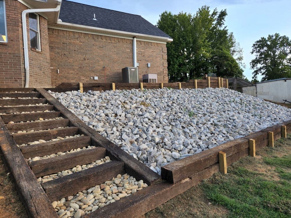 Landscape featuring a rock-filled slope with wooden steps leading to a brick house.