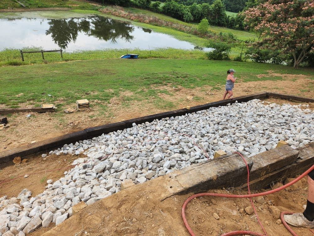 Child playing near a gravel area by a pond, with trees and greenery in the background.