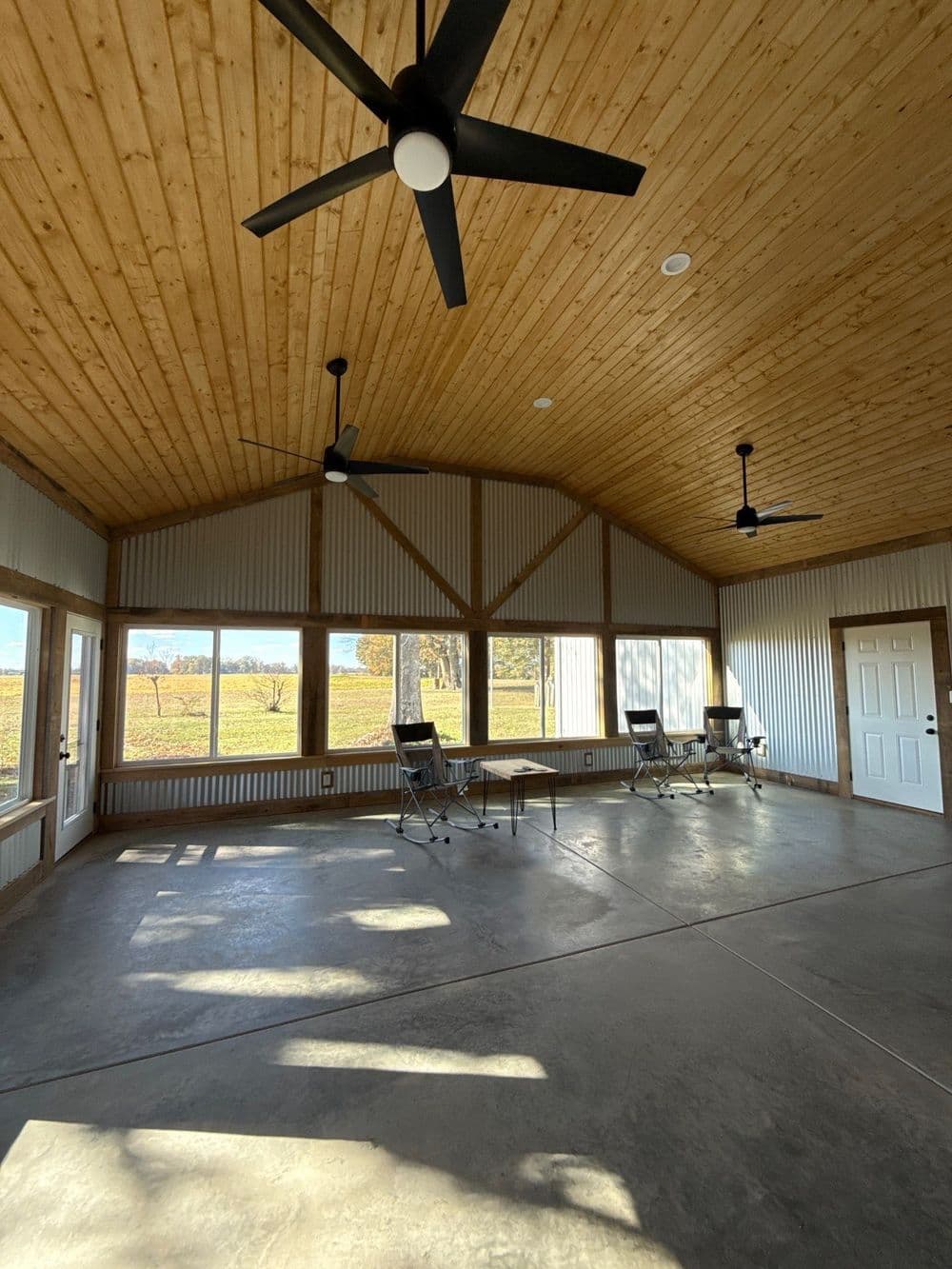 Spacious indoor room with wooden ceiling, large windows, and ceiling fans overlooking nature.